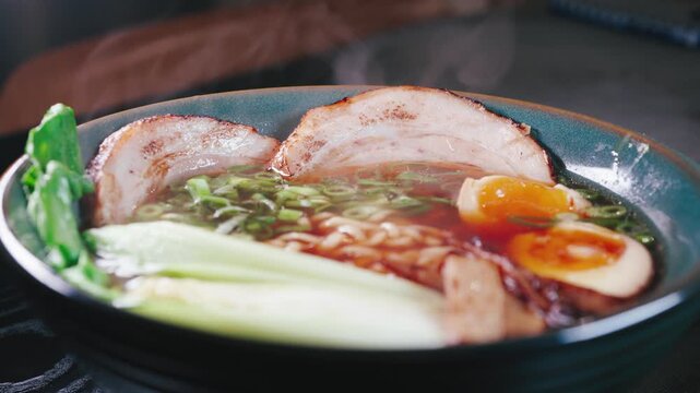 Close-up video of steaming ramen made with instant noodles, topped with tender chashu pork belly, soft boiled egg, fresh bok choy and bamboo shoots in a rich soy pork broth. Hot vapor rising from the 