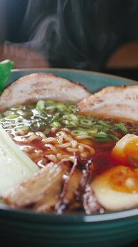 Close-up video of steaming ramen made with instant noodles, topped with tender chashu pork belly, soft boiled egg, fresh bok choy and bamboo shoots in a rich soy pork broth. Hot vapor rising from the 
