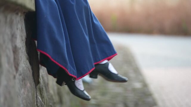 Woman in traditional Latvian attire sits on a stone wall, showcasing her black shoes and blue skirt with red trim in a natural setting
