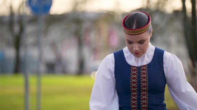 Latvian woman in traditional attire stands gracefully outdoors, showcasing cultural heritage with vibrant colors and intricate patterns in a park setting