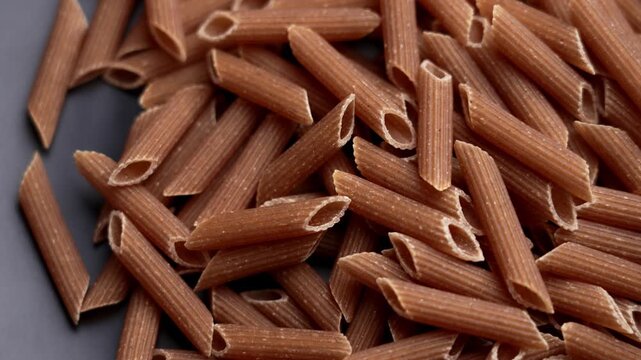 Pile of integral whole wheat penne rigate pasta on black ceramic plate. Dry uncooked tubular macaroni with ridged surface. Italian food ingredient. Rotation. Top view