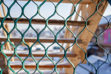 Fototapeta premium Green patterned fence in front of wooden stairs with snow-covered ground and blurred background of vehicles and buildings