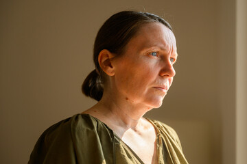 Fototapeta premium Pensive mature woman illuminated by soft window light, gazing outside with introspective expression, emotional lifestyle portrait in a calm home interior.