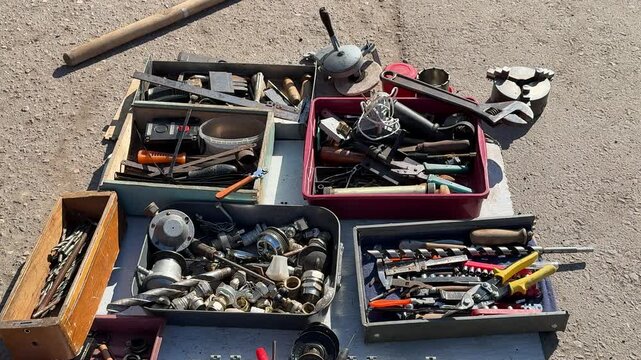 Tools for sale on asphalt showing assorted pliers, wrenches, screwdrivers and sockets in open boxes jars of screws and rusty.