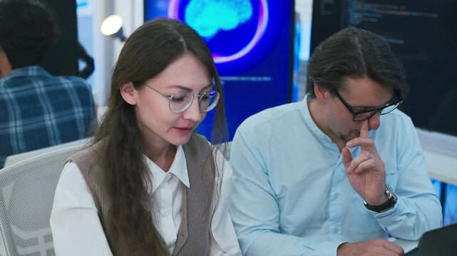 collaborative AI development team in modern tech office: young male developer coding on laptop, female team leader in light blue blazer pointing at code and neural network visualization	
