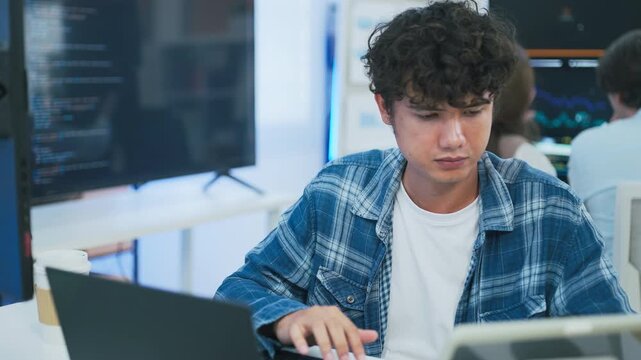 focused young male developer with curly hair in blue plaid shirt intensely codes on laptop in modern AI office, surrounded by multiple monitors displaying code neural networks and data graphs	