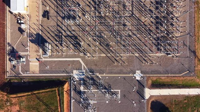 Overhead aerial view of a large electrical substation with transformers, switchgear, and power lines casting long shadows across the gravel yard, surrounded by industrial facilities