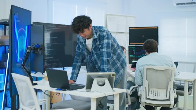 focused young male developer with curly hair in blue plaid shirt intensely codes on laptop in modern AI office, surrounded by multiple monitors displaying code neural networks and data graphs	