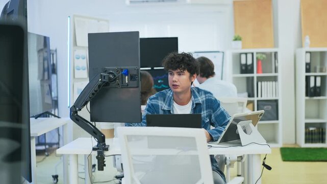 focused young male developer with curly hair in blue plaid shirt intensely codes on laptop in modern AI office, surrounded by multiple monitors displaying code neural networks and data graphs	