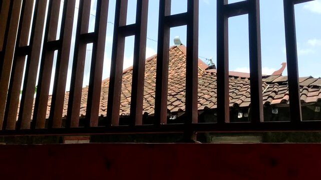 Traditional Indonesian house featuring classic terracotta clay roof tiles, captured from behind a dark iron vertical slat fence. Authentic Javanese architectural elements.