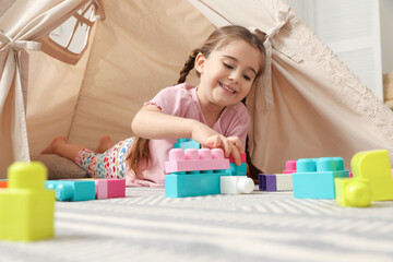 Little girl playing with building blocks near toy wigwam in playroom, low angle view © New Africa