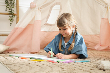 Little girl drawing in sketchbook near toy wigwam at home © New Africa