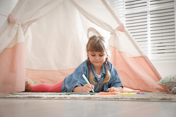 Little girl drawing in sketchbook near toy wigwam at home © New Africa
