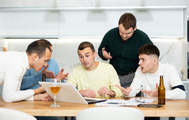 Fototapeta premium Concerned group of friends offering support to upset man receiving bad news, gathering together around laptop at home table during informal meeting with beer