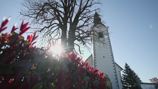 Slow motion video of Church tower in Grosuplje rising beside a leafless tree, captured from a low angle with sun flare through red foliage, creating a dramatic perspective and serene atmosphere on a