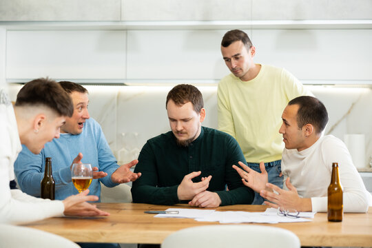 Man appearing upset as reading document, while friends gathering around, offering support and advices during friendly meeting at home kitchen..