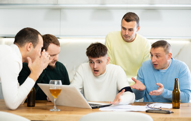 Fototapeta premium Concerned group of friends offering support to upset man receiving bad news, gathering together around laptop at home table during informal meeting with beer
