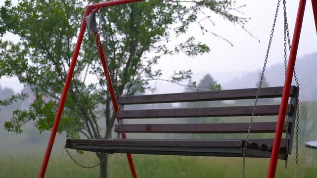 Empty wooden swing gently swaying in the summer rain. Sad and lonely empty wooden swing with a red frame gently swaying in the backyard during a moody summer rain shower