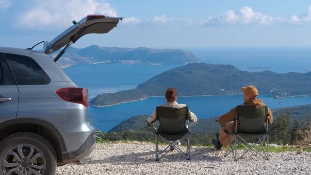 Couple enjoying scenic view from mountaintop with car. Tourists relaxing in camping chairs, admiring a breathtaking view of the sea and islands from a mountain peak, with their car parked nearby