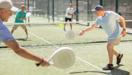Obraz premium Adult man and elderly man doubles playing against two men in padel tennis on court