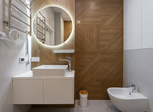 Modern bathroom interior with round LED mirror, wooden geometric wall tiles, white vanity sink and minimalist sanitary ware in stylish contemporary apartment design