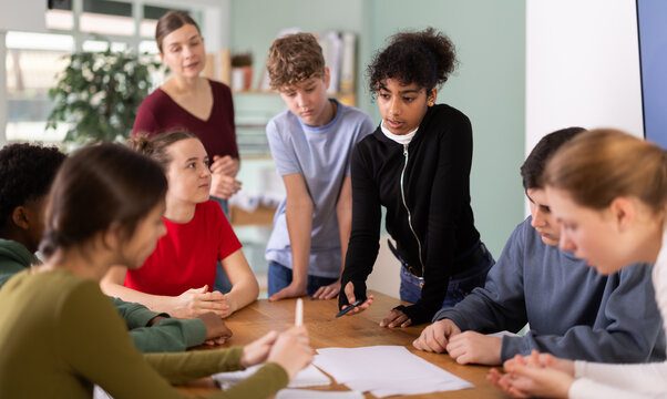 Group of teenagers and students discuss the task at the table, they exchange opinions and theories against the background of teacher in classroom. Schoolchildren look at the paper and solve the task