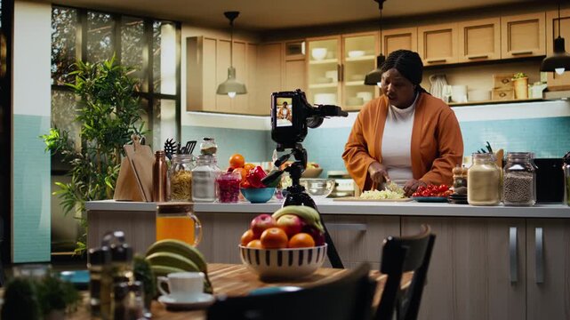 Woman influencer chef recording a food vlog and cutting iceberg salad, kitchen studio with natural light. African American content creator explains the cooking process and preparation.