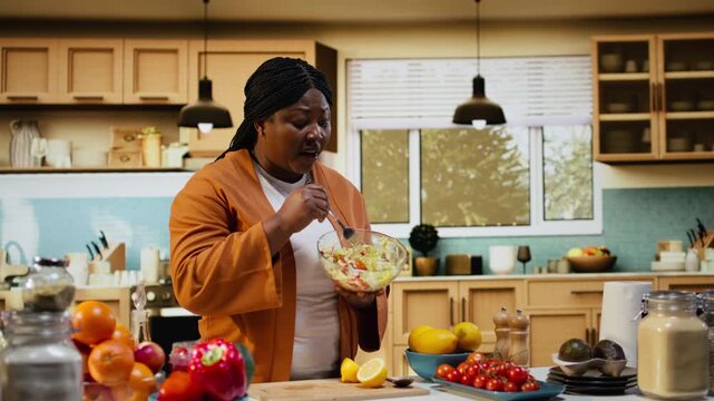 Black food vlogger mixing vegetables for a salad recipe in bowl, creating lifestyle cooking video in the kitchen. Home chef organizing ingredients on the counter, recipe tutorial and meal prep.