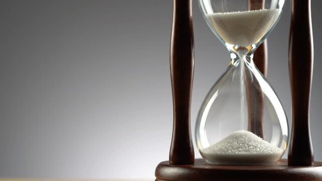 White sand flows through a traditional wooden hourglass against a grey background, symbolizing the passage of time and deadlines.