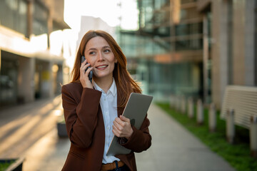Businesswoman talking on smartphone walking with laptop outside office