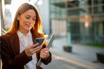 Businesswoman checking phone holding laptop smiling outdoors