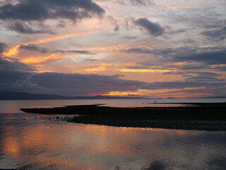 Obraz premium Serene coastal sunset over calm waters and silhouetted hills. Dramatic orange and purple clouds reflecting on the tide at dusk. Peaceful evening landscape in New Zealand with a lone fisherman.