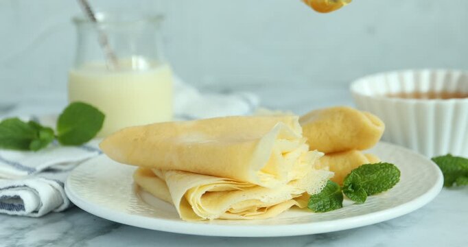 Woman dripping honey onto tasty crepes at white marble table, closeup