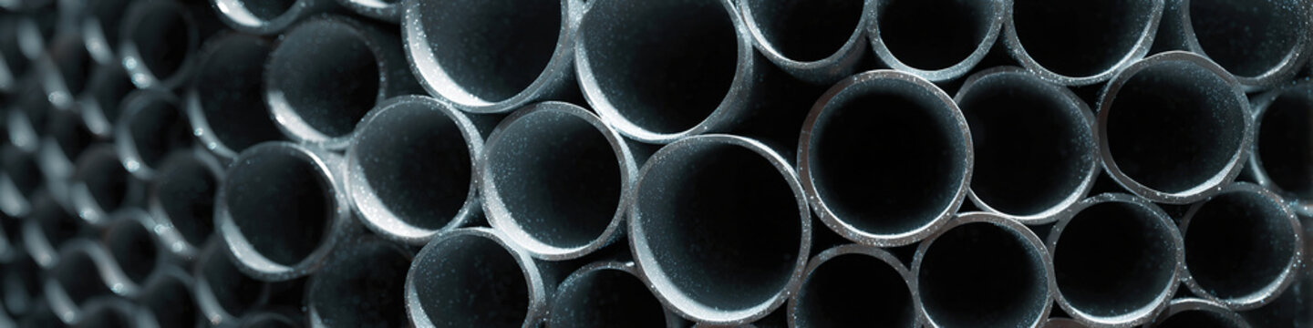 Dark metal pipes stack tightly, circular openings facing forward. Industrial texture shows wear, depth, and uniform shape. Strong contrast highlights edges, inner hollows, and cylindrical form