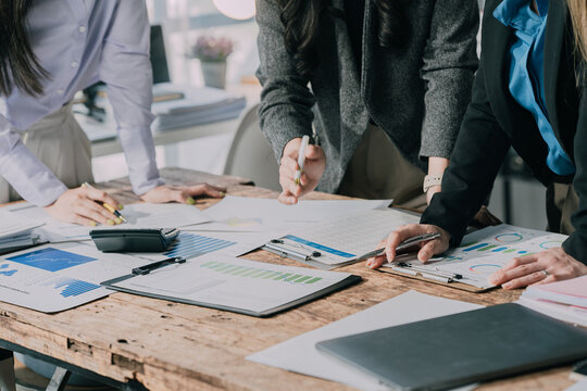 A group of business people are discussing business or projects. They are holding pens and pointing to printed documents containing charts and graphs, likely performance data or reports, indicating col