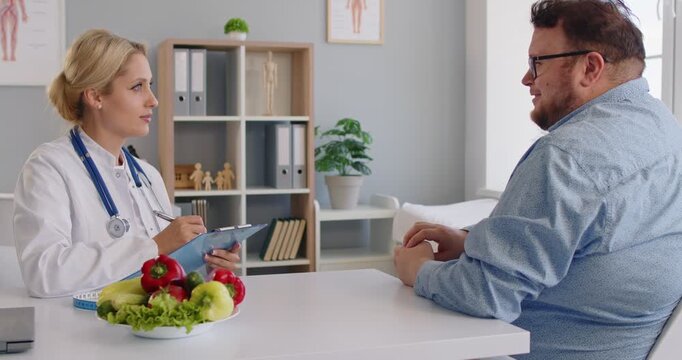 Nutritionist leads a healthcare clinic consultation, reviewing diet notes with a fat overweight patient and discussing obesity risks, and advice for wellness. Doctor guides patient on diet today.