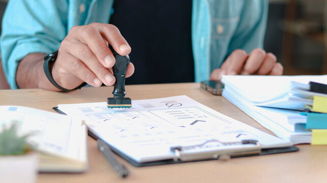 Close-up of business professional stamping approval on financial report documents at desk. Concept of authorization, verification, paperwork, compliance, and workflow process.