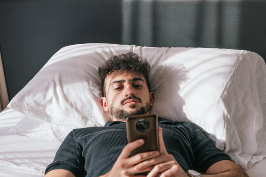 Young man lying in bed, using his mobile phone to scroll through social media or news in the morning