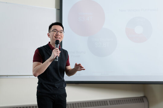 A smiling Asian man wearing glasses holds a microphone while gesturing during a presentation in front of a chart, ideal for business pitch, school presentation, or public speaking concepts.