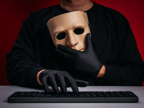 Cybercrime and online fraud concept with anonymous person wear gloves, holding mask and using computer keyboard. Hacker attack, phishing scam, identity theft, deepfake threat and cybersecurity risk.