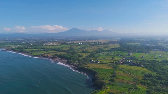 Stunning aerial view of Bali coastline featuring deep blue ocean waves, lush green rice fields, and the majestic Mountain Batu karu volcano under a clear morning sky in Indonesia.