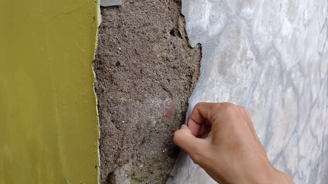 Wide shot of Hand peels away crumbling concrete wall layer. Texture shows rough, gritty surface beneath. Grayish hue suggests age or weather exposure. Action implies renovation, repair, or demolition 