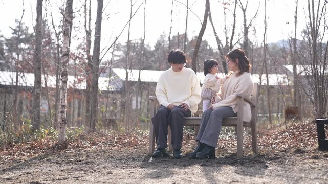 Soft sunlight bathes a quiet countryside scene. Family sits together on a simple wooden bench. Their calm expressions suggest peaceful, intimate moments. Autumn leaves cover the ground around them