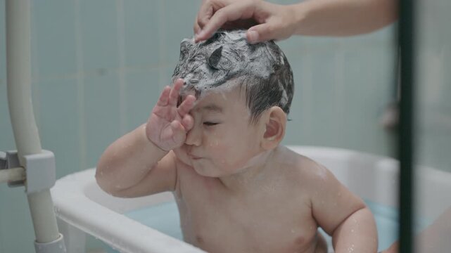 Adorable Asian baby boy taking a bath in a small tub with soap bubbles on head. Looking at camera with funny expression.