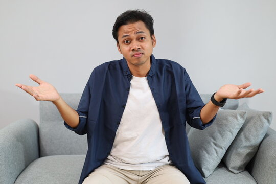 An young man sits on a sofa at home while shrugging his shoulders and opening his palms, with a confused, questioning expression, looking at the camera.