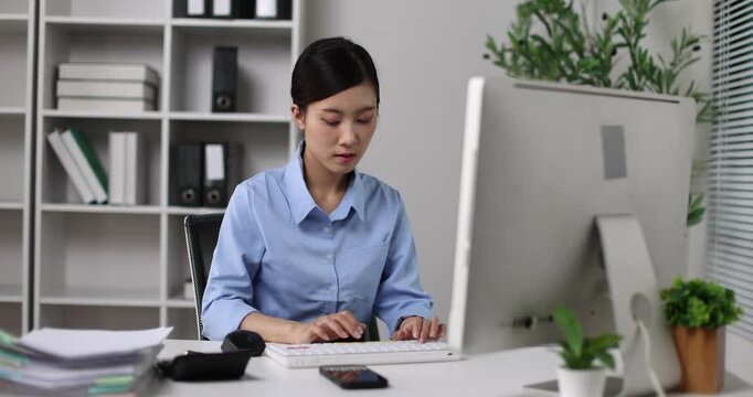 Asian businesswoman working diligently at her desk in a contemporary office setting, performing data entry and managing tasks on a desktop computer, focusing on productivity and professional work
