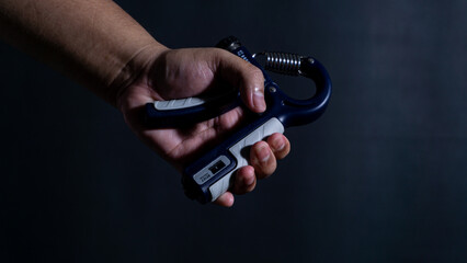 Fototapeta premium A male hand tightly clamps down on a blue and grey hand strengthener in front of a black background, indicating an intense grip training session. The single light source creates heavy