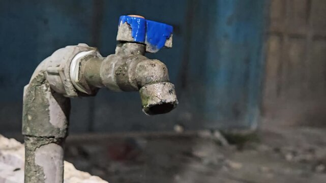 Close-up of an old, corroded water faucet slowly dripping in a dimly lit, grungy environment. The dirty tap with a broken blue handle highlights severe infrastructure decay