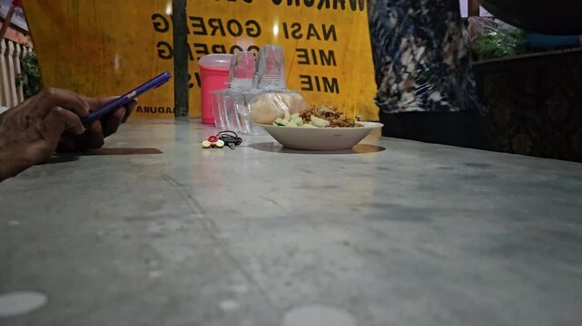 Close-up of a person's hands holding and using a blue smartphone while sitting at a street food stall counter at night. In the background, a vendor is actively cooking in a wok.