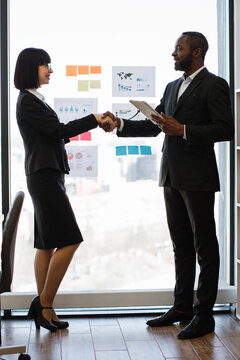 A professional handshake between a businesswoman and an African American businessman in an office setting with charts on the wall